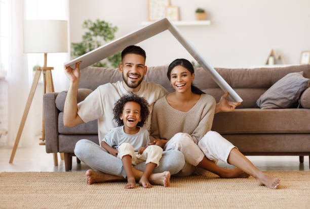Cheerful parents with child smiling and keeping roof mockup over heads while sitting on floor in cozy living room during relocation